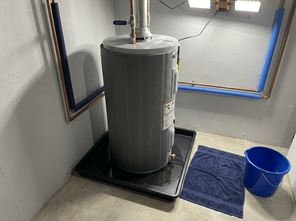Basement Water Heater Leaking At The Bottom With Water Pooling On The Floor, A Drain Pan In Place, And Emergency Items Nearby Like A Towel And Bucket.